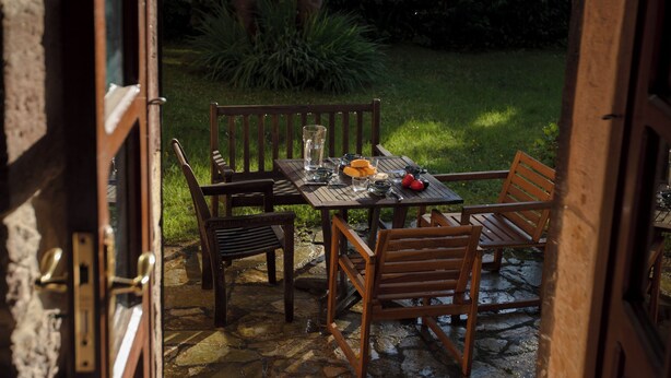 Un patio al aire libre con una mesa y sillas de piedra, rodeado de abundante vegetación y una vista de un edificio al fondo.