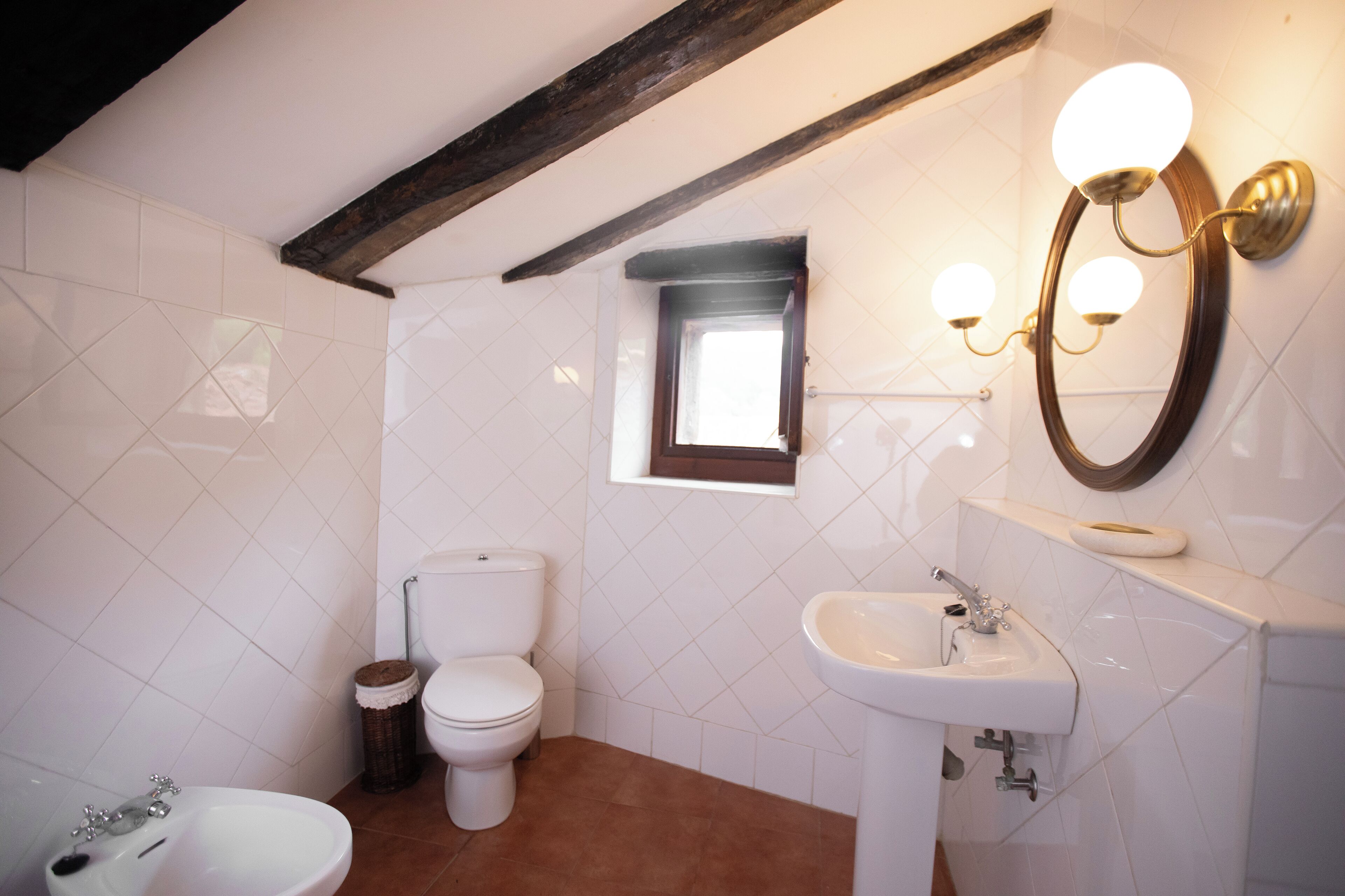 Een badkamer met toilet, wastafel en bidet. Er is een raam boven het toilet en een spiegel met verlichting boven de wastafel.