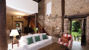 Un interior rústico de piedra con un sofá blanco, cojines verdes, una mesa de centro de madera y un sillón a rayas.
