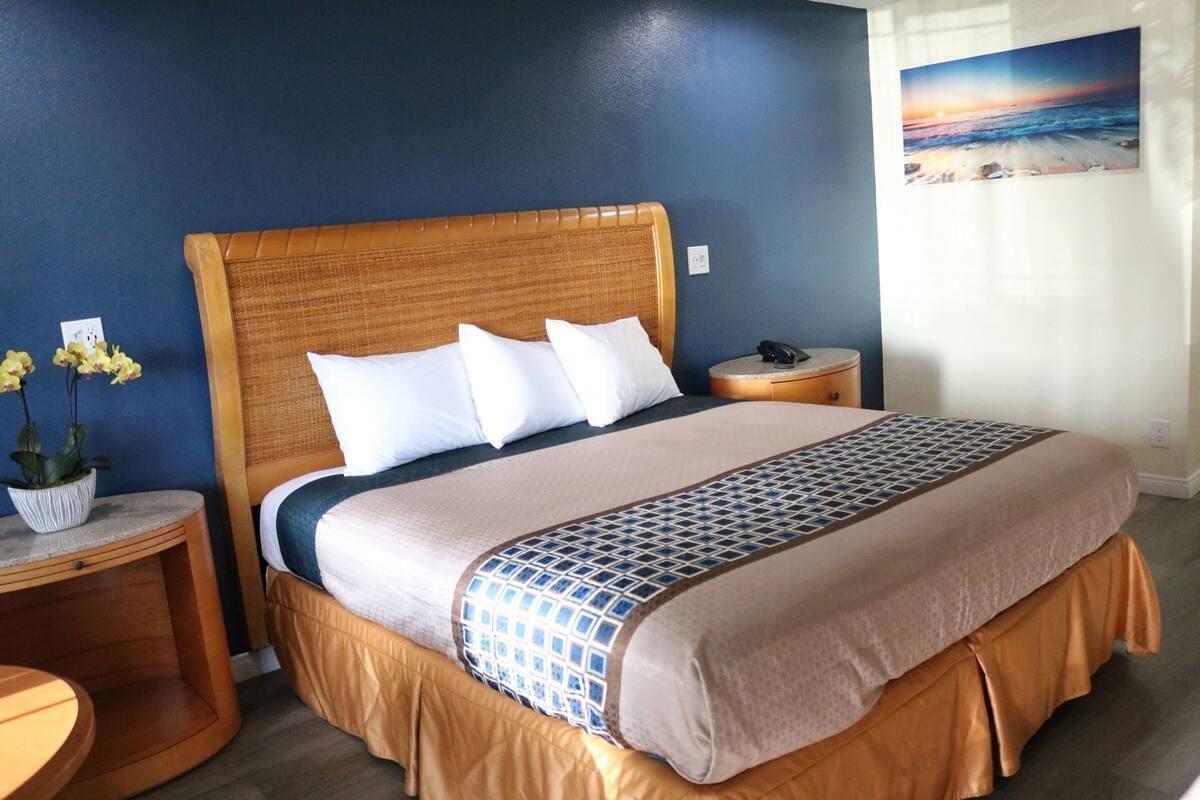 A hotel room with a large bed, a nightstand, a bedside table, a potted plant, and a painting of a beach scene.