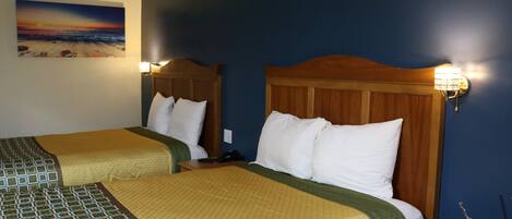 A hotel room with two beds, each with a wooden headboard and white bedding. There is a painting of a beach scene on the wall, and a nightstand with a lamp between the beds.