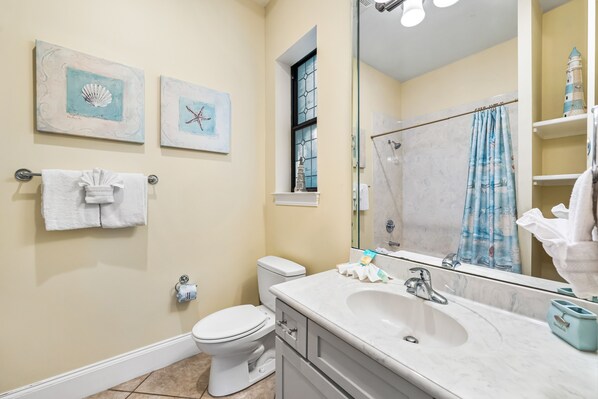 A bathroom with a toilet, sink, and a large mirror. There are two framed pictures on the wall, a towel rack with a towel, and a shower curtain.