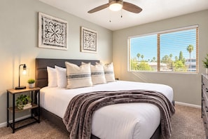 A bedroom with a bed, nightstand, lamp, ceiling fan, window with blinds, and two wall-mounted decorative pieces. - Walk to Old Town | 2 King Beds + 4 Heated Pools (Scottsdale)