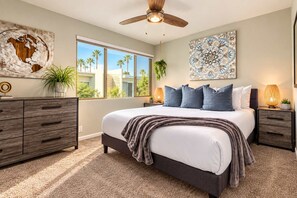 A bedroom with a large bed, a ceiling fan, a window with a view of palm trees, and a decorative wall piece.