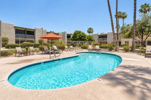 A swimming pool with lounge chairs and an umbrella, surrounded by a fence and palm trees.