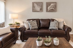 A brown leather sofa with patterned cushions, a wooden coffee table with potted plants, and two wall-mounted artwork pieces. - Walk to Old Town | 2 King Beds + 4 Heated Pools (Scottsdale)