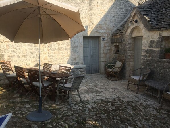 An outdoor patio with stone flooring, a wooden table with chairs, a large umbrella, and wicker chairs. - Relaxing Trullo -
Original and comfortable trullo in Valle d'Itria Puglia (Locorotondo, Bari)