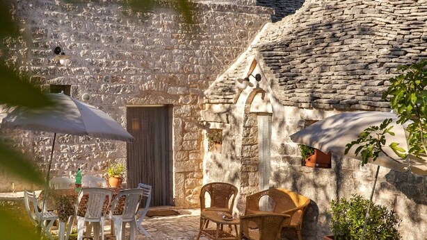 Un edificio de piedra con fachada rústica, zona de estar exterior con sillas de plástico blanco y mesas de mimbre, y una planta en maceta.