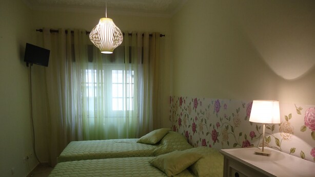 A bedroom with two beds, a bedside table with a lamp, a TV on the wall, and a window with sheer curtains.