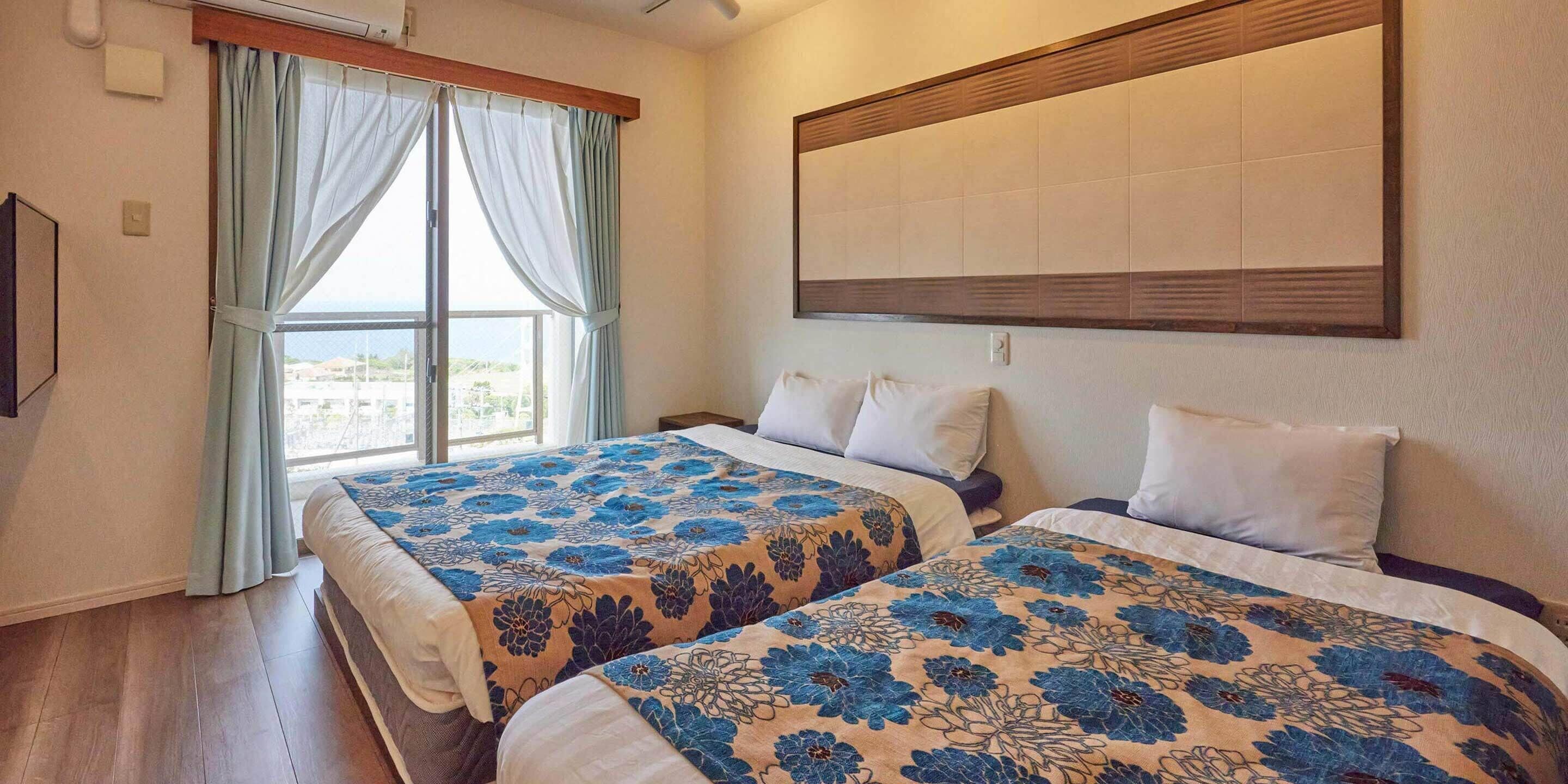 Une chambre d’hôtel avec deux lits, une grande fenêtre avec des rideaux, un téléviseur fixé au mur et une tête de lit en bois.
