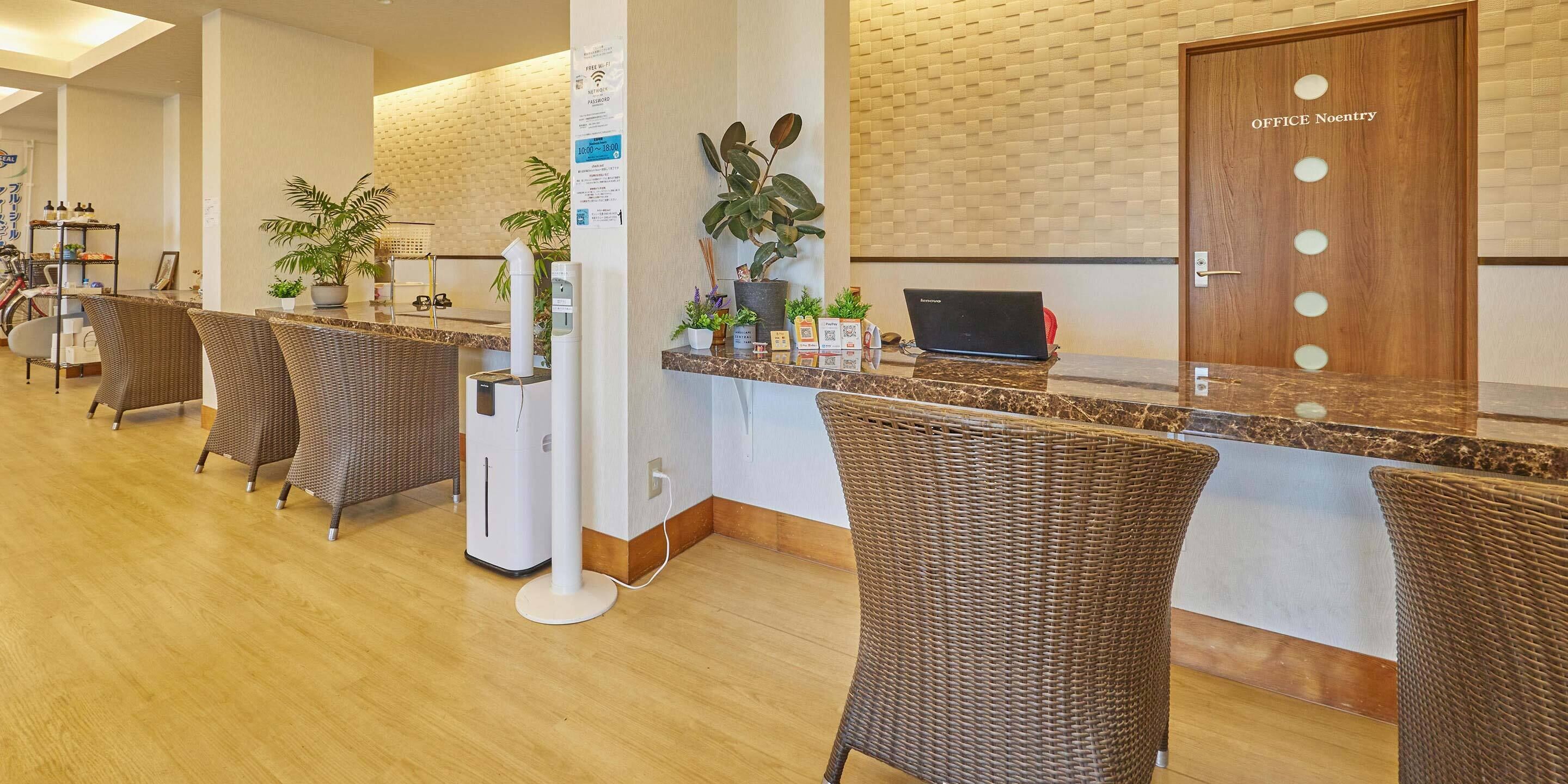 A hotel reception area with a wooden reception desk, wicker chairs, potted plants, and a laptop on the desk.