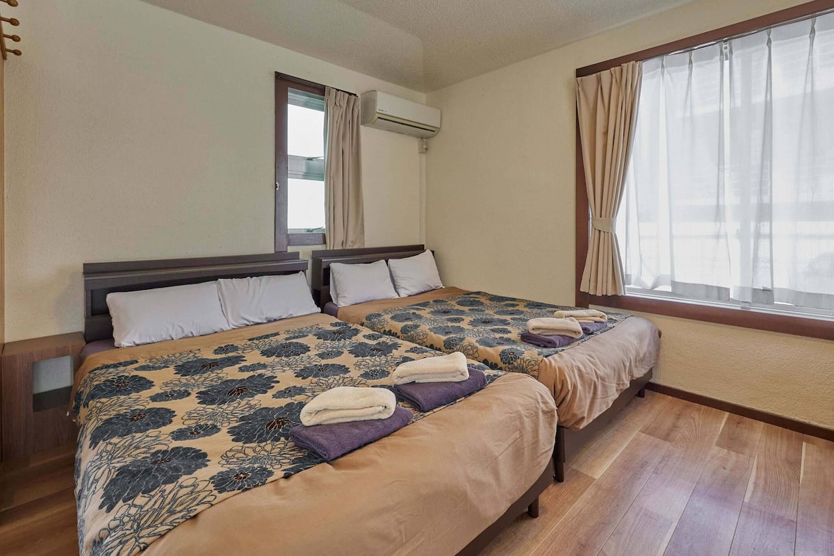 A hotel room with two beds, a window with curtains, an air conditioning unit, and wooden flooring.