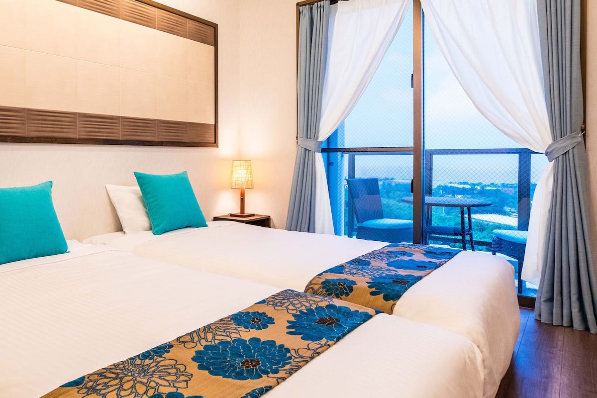 A hotel room with two beds, a large window with curtains, a view of the city, and a nightstand with a lamp.