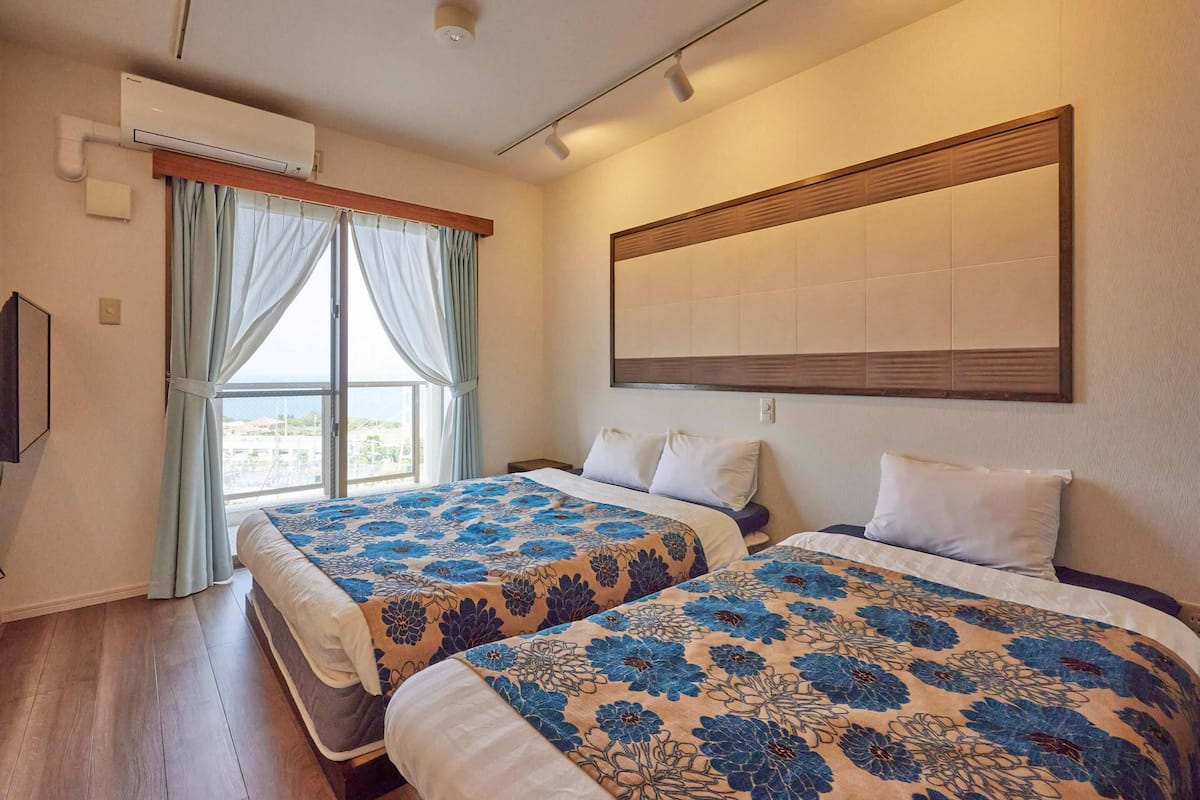 A hotel room with two beds, a large window with curtains, an air conditioning unit, and a mounted artwork on the wall.