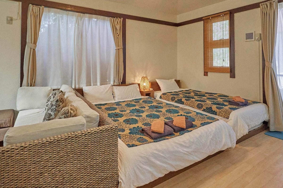 A hotel room with two beds, a sofa, a small table, and large windows with curtains.
