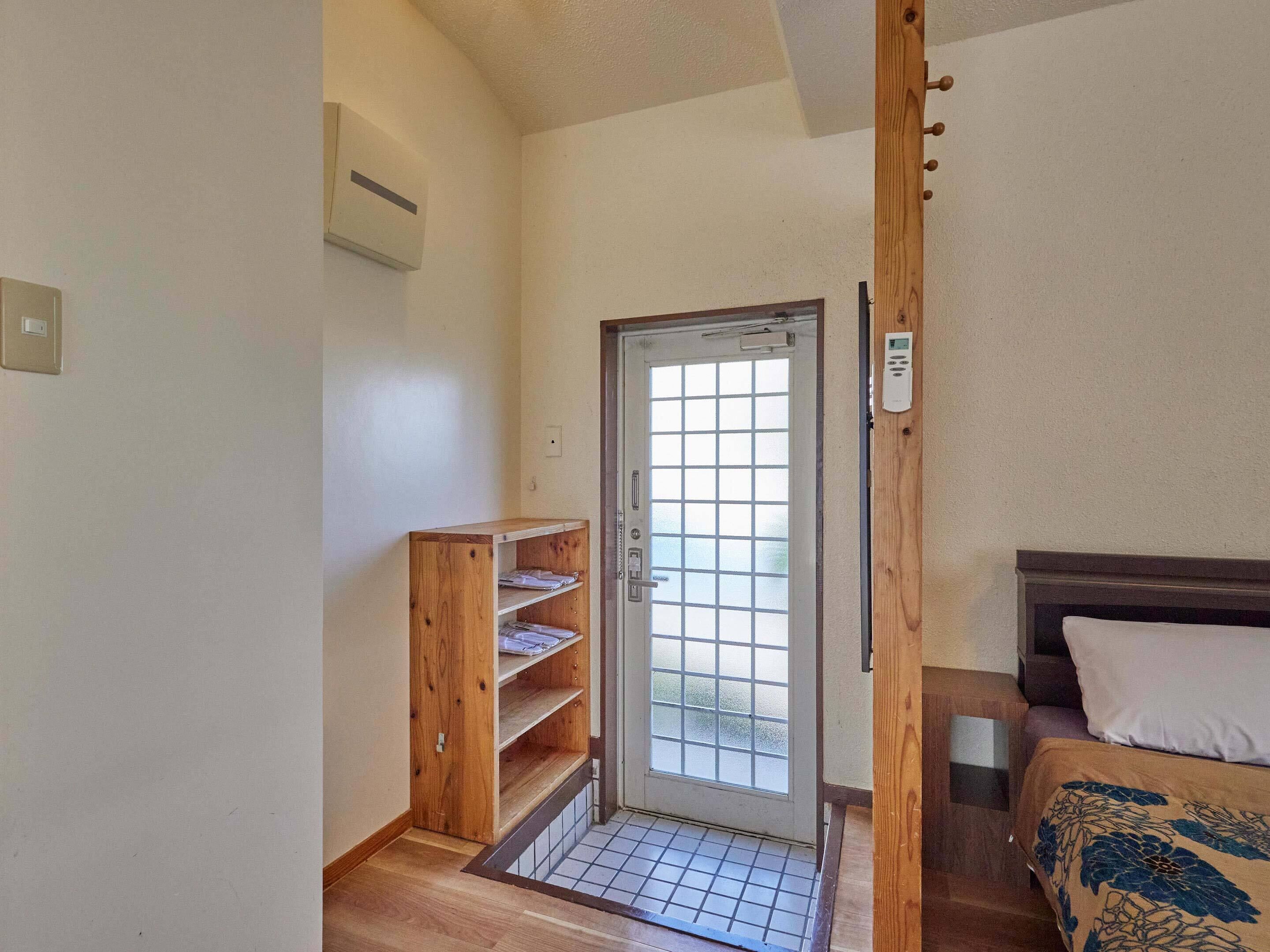 A room with a wooden door, a small shelf unit, a bed with a patterned bedspread, and a thermostat on the wall.