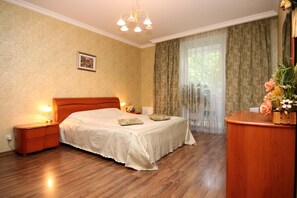 Un dormitorio con una cama de madera, mesitas de noche, una araña, un cuadro enmarcado en la pared y un escritorio de madera con un florero.