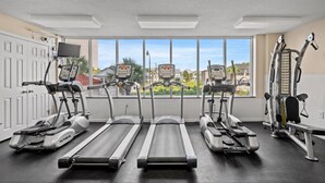 A gym with treadmills, ellipticals, and weight machines, large windows with a view of houses, and a door with a security camera. - Cozy Corner Unit, Jacuzzi Tub and Recliner, Lazy River | Crescent Shores - S 312 (North Myrtle Beach)