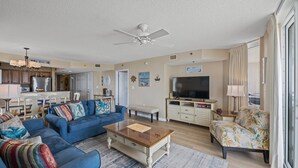 A spacious living room with a blue sofa, a coffee table, and a television. There is a kitchen area with cabinets and a refrigerator. - Amazing Oceanfront Condo, Indoor and Outdoor Pools | Crescent Shores - S 912 (North Myrtle Beach)