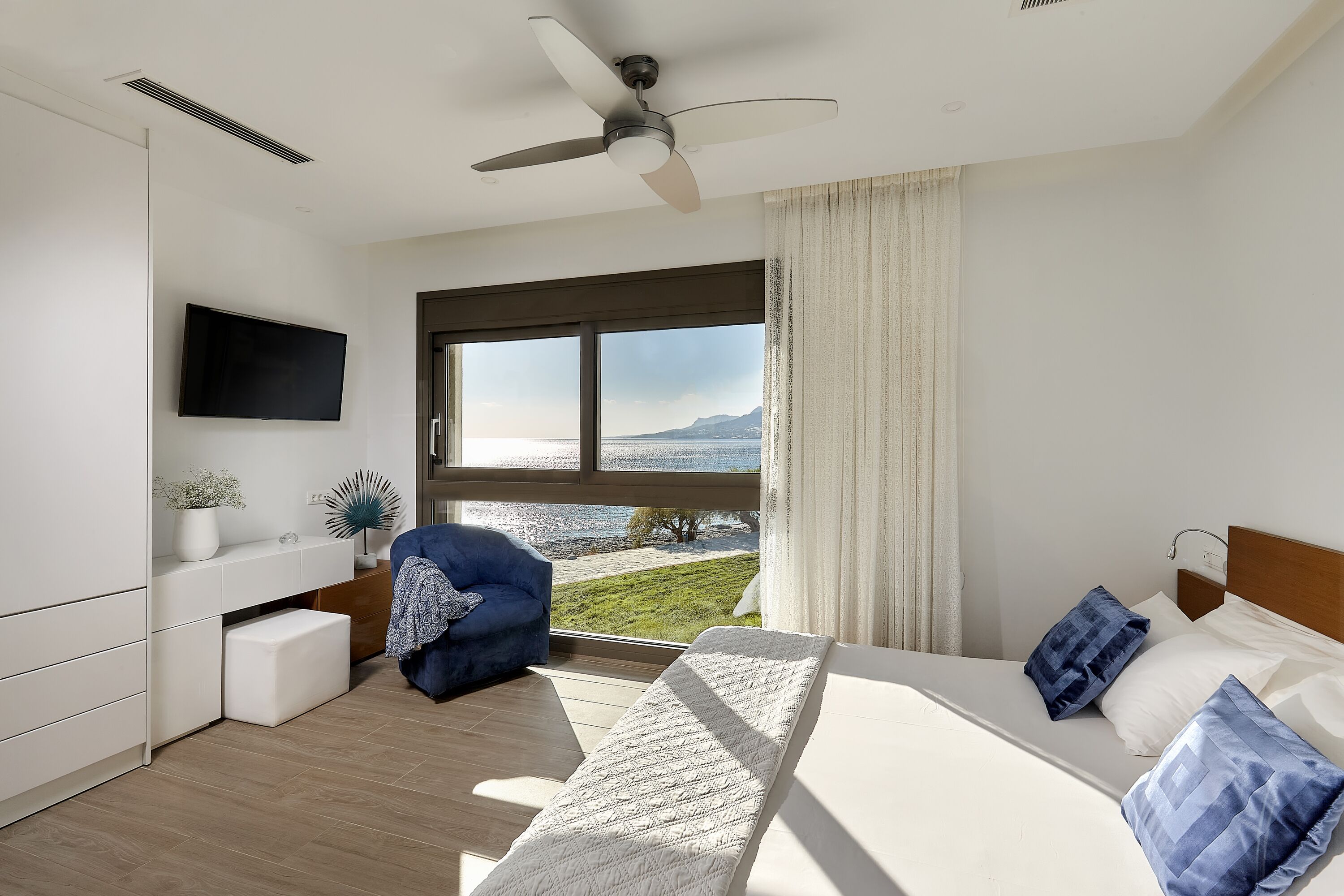 A modern hotel room with a bed, a desk, a chair, a TV, and a large window with a view of the sea and mountains.