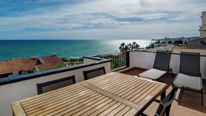 Une terrasse dotée de tables et de chaises en bois, offrant une vue sur la côte, avec des bâtiments et un ciel dégagé.