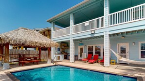 A two-story building with a pool, a thatched-roof structure, and red lounge chairs. - Private Heated Pool, Golf Cart for Entire Stay, Game Room, Pet Friendly (Port Aransas)