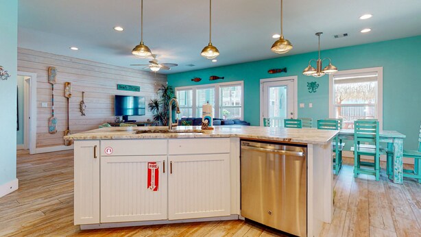 A kitchen with a large island, white cabinets, and a dishwasher. There are pendant lights, a dining area with a table and chairs, and a view of the outside through large windows. - Private Heated Pool, Golf Cart for Entire Stay, Game Room, Pet Friendly (Port Aransas)