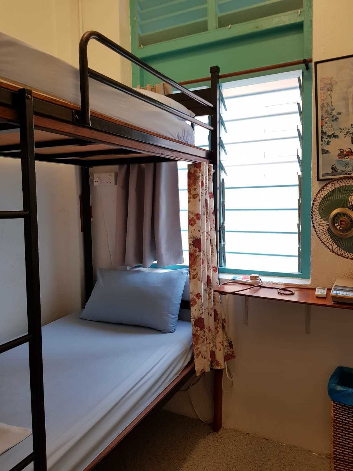 Uma cama de beliche com escrivaninha e cadeira, uma janela com cortinas e um ventilador fixado na parede.
