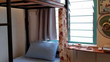 Uma cama de beliche com escrivaninha e cadeira, uma janela com cortinas e um ventilador fixado na parede.
