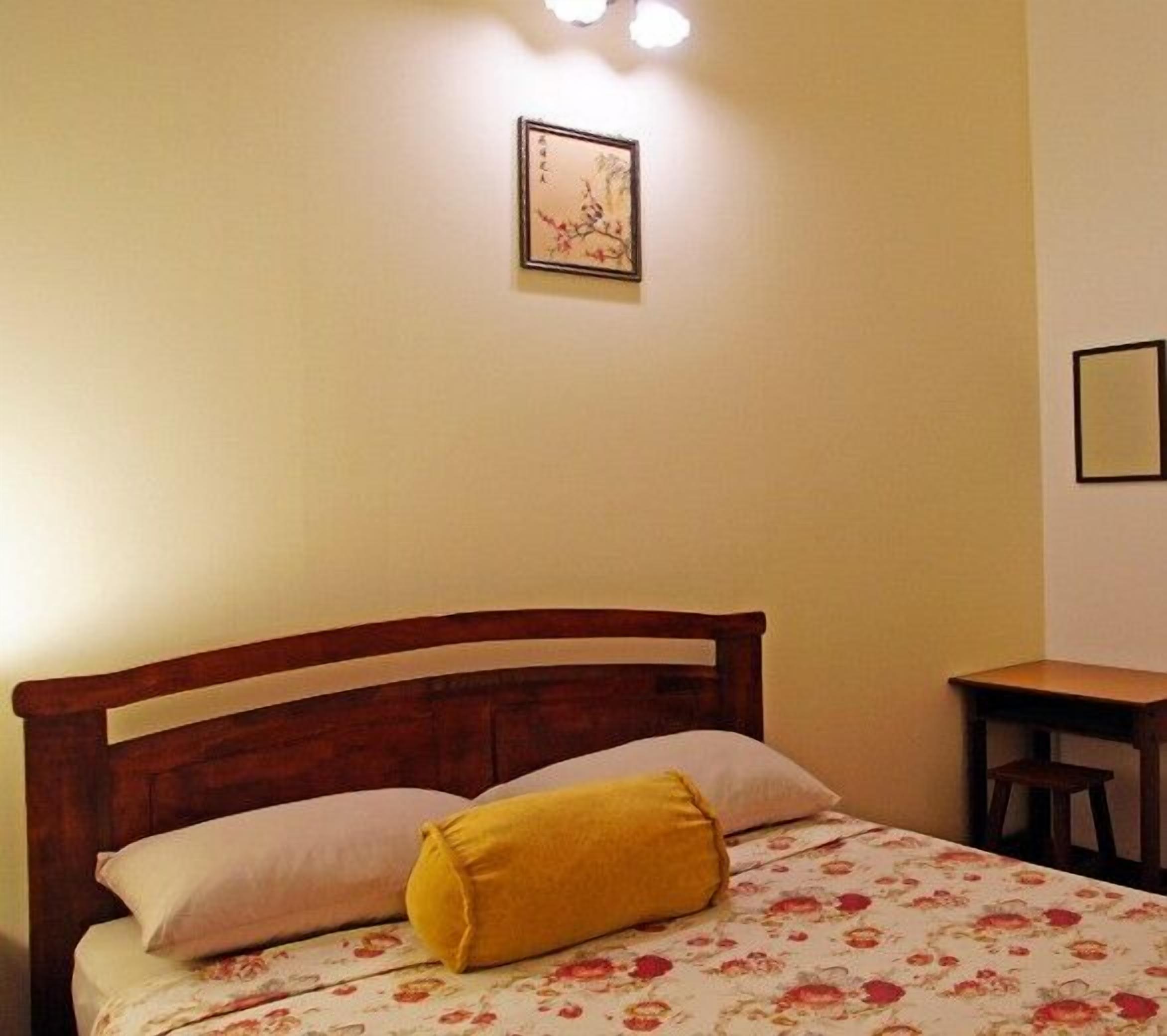 Uma cama com cabeceira de madeira, duas almofadas e um cobreleito floral. Há uma pequena mesa à direita e um quadro na parede acima da cama.
