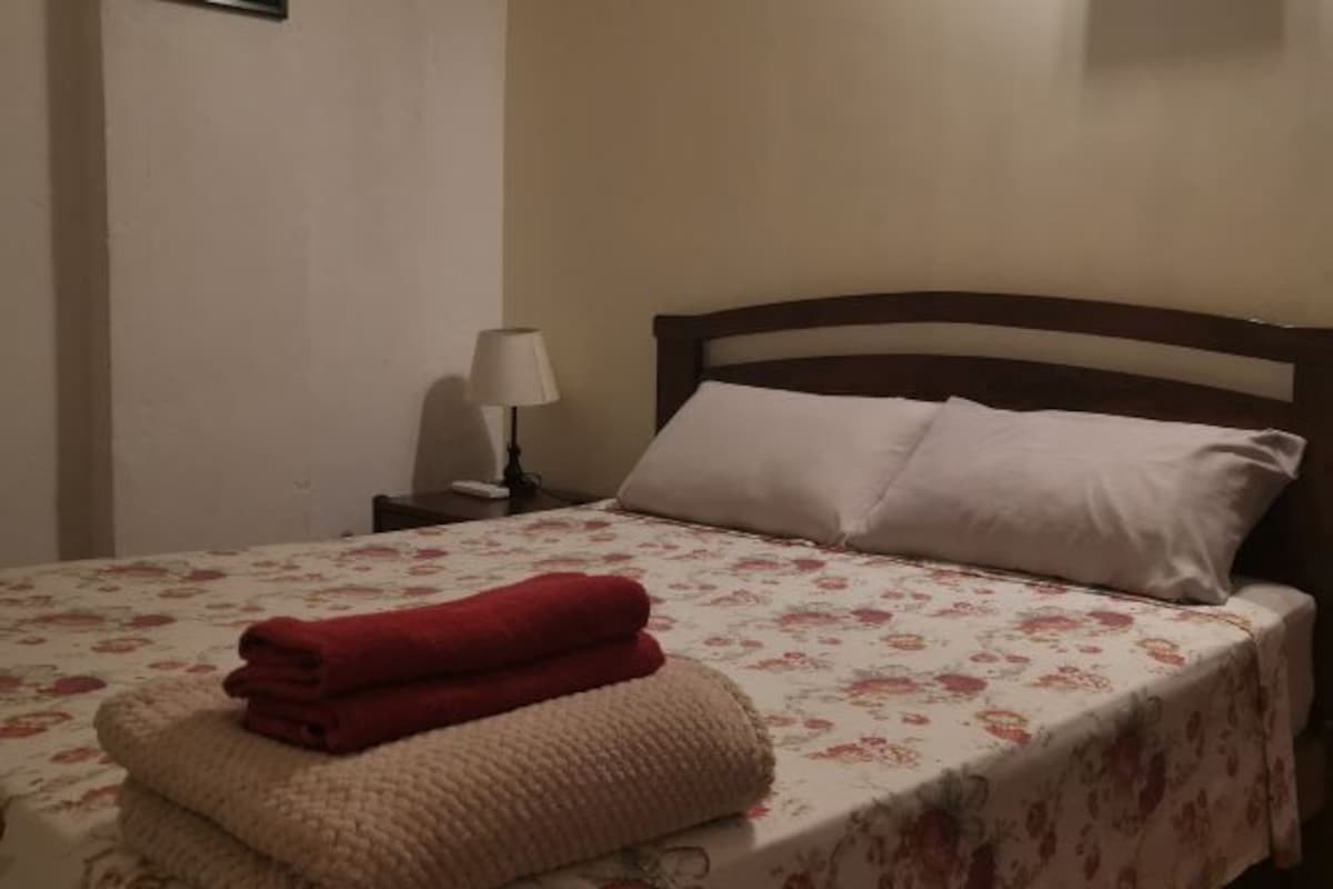 A bed with a floral bedspread, two pillows, a nightstand with a lamp, and folded towels.