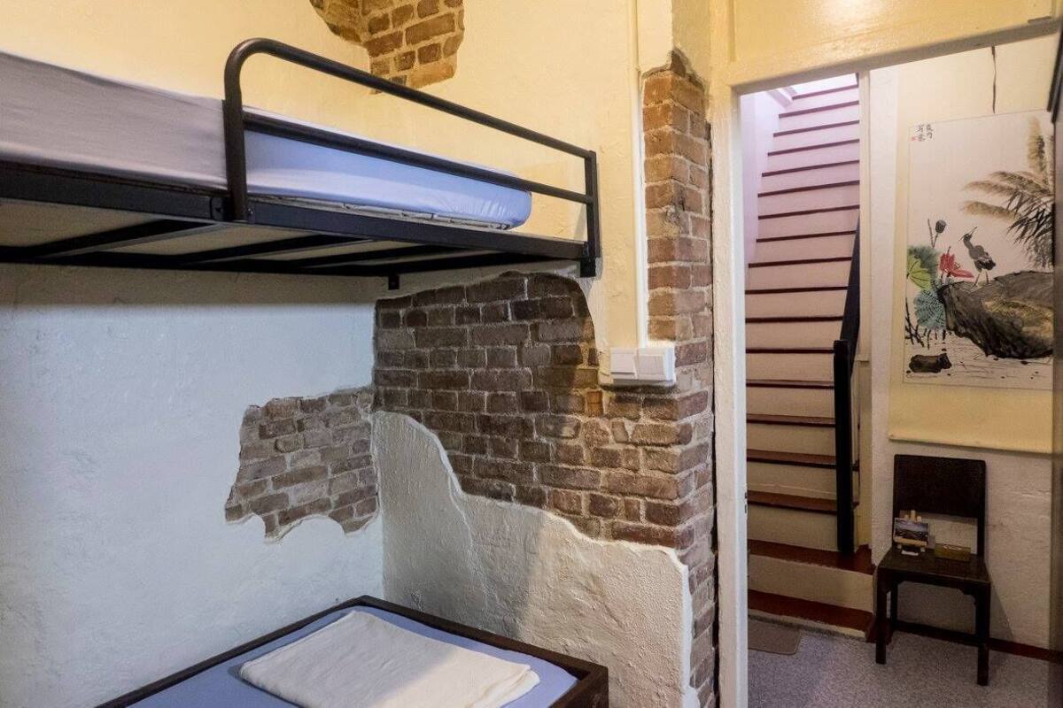 A bunk bed with a mattress, a brick wall, a small table with a white cloth, and a painting on the wall.