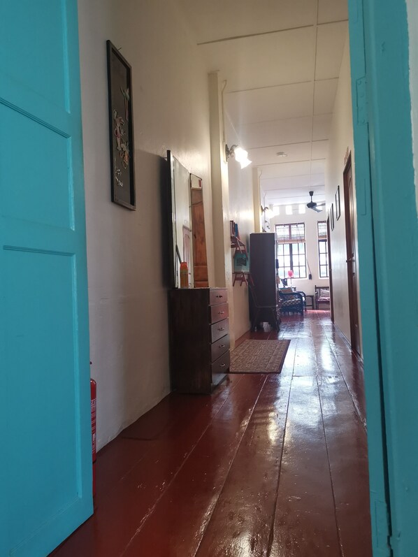 Un pasillo con suelo de madera, un espejo y un aparador. Se ven ventanas y un ventilador de techo al fondo.
