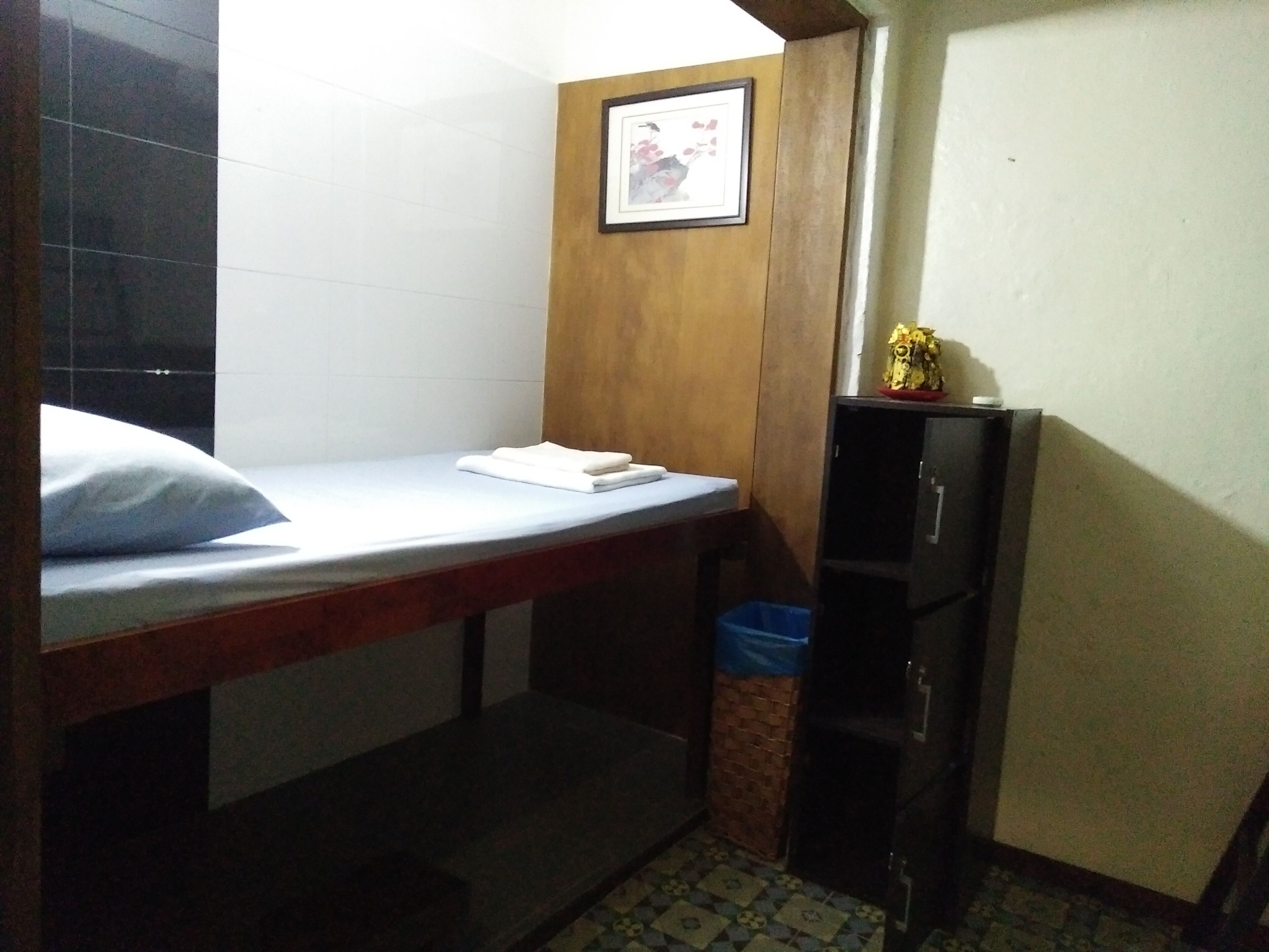 Una habitación pequeña y estrecha con una cama individual, un armario de madera y un cubo de basura.