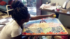 Una persona sentada a una mesa con cubierta de mosaico, una silla y una planta en maceta al fondo.