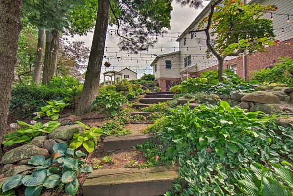 Un chemin de jardin mène à un bâtiment à la façade en briques, entouré d’une végétation luxuriante et de marches en pierre.