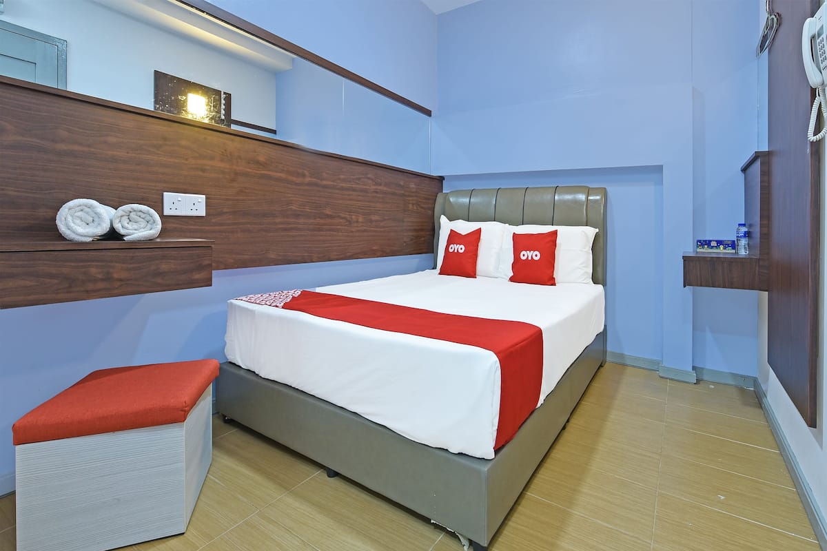 Un lit bien fait, avec une tête de lit grise, une couvre-lit rouge et blanche et des coussins assortis. On y trouve une petite table de chevet blanche avec un coussin rouge. Les murs de la pièce sont bleu clair, un mur est mis en valeur avec du bois, et le sol est en bois.