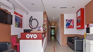 Zona de recepción de un hotel con mostrador de recepción identificado como 'OYO', un televisor montado en la pared, una silla roja, un sofá negro y un pasillo que conduce a una habitación.