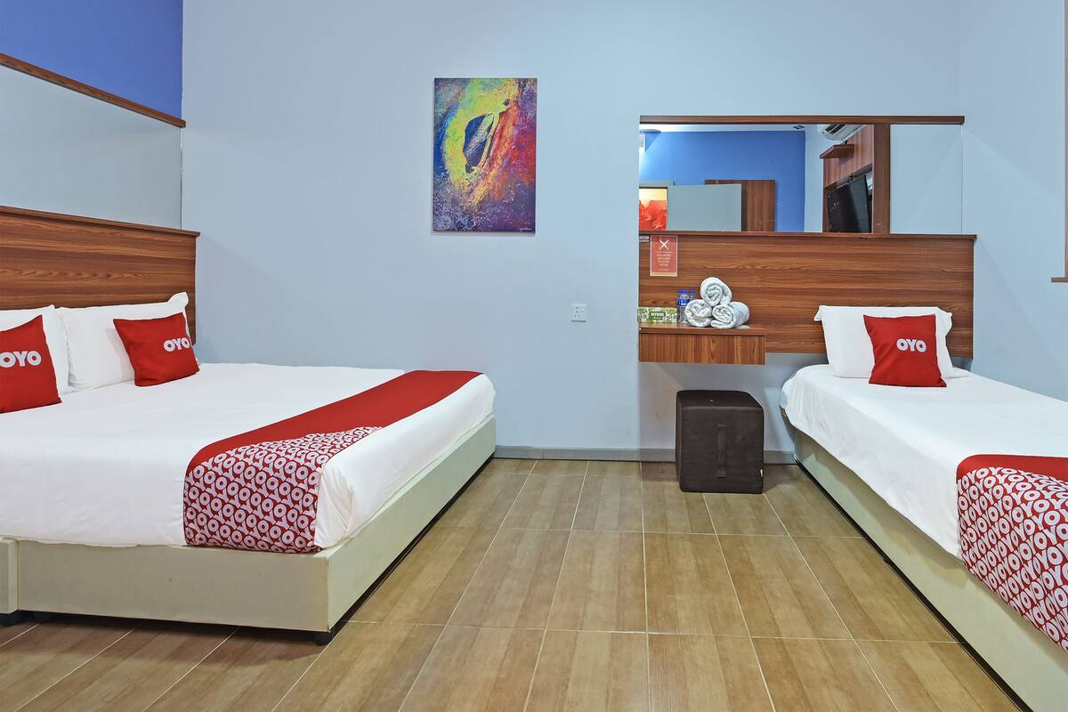 Une chambre d’hôtel avec deux lits, chacun recouvert d’une courtepointe rouge et blanche et agrémenté d’oreillers, une tête de lit en bois, une petite table de chevet avec des articles de toilette et une peinture abstraite aux couleurs vives accrochée au mur.