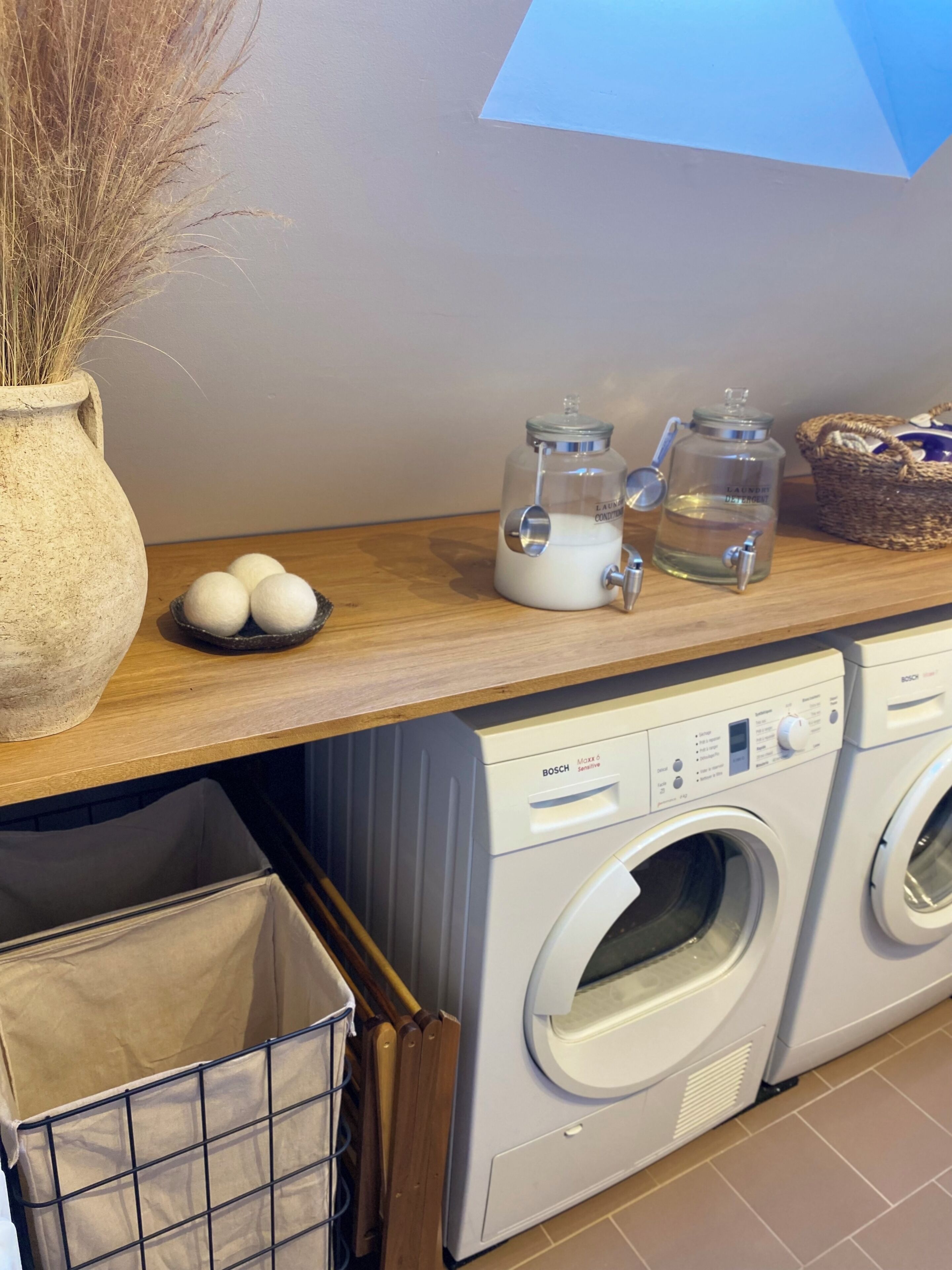 Een wasruimte met een wasmachine, een droogmachine, een vaas met gedroogde planten en een mand.