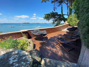 A wooden deck with a rocking chair and lounge chairs overlooking a body of water with boats.