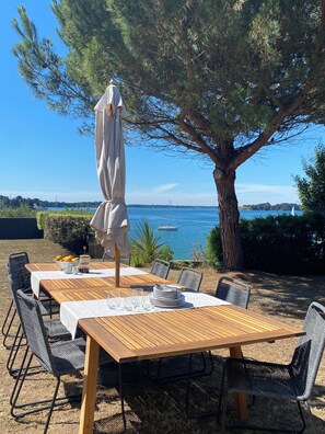 An outdoor dining area with a table set for four, surrounded by chairs, under a large tree, with a view of the sea and boats. - "Heaven's door", exceptional villa feet in the water, direct access to the sea (Sarzeau)