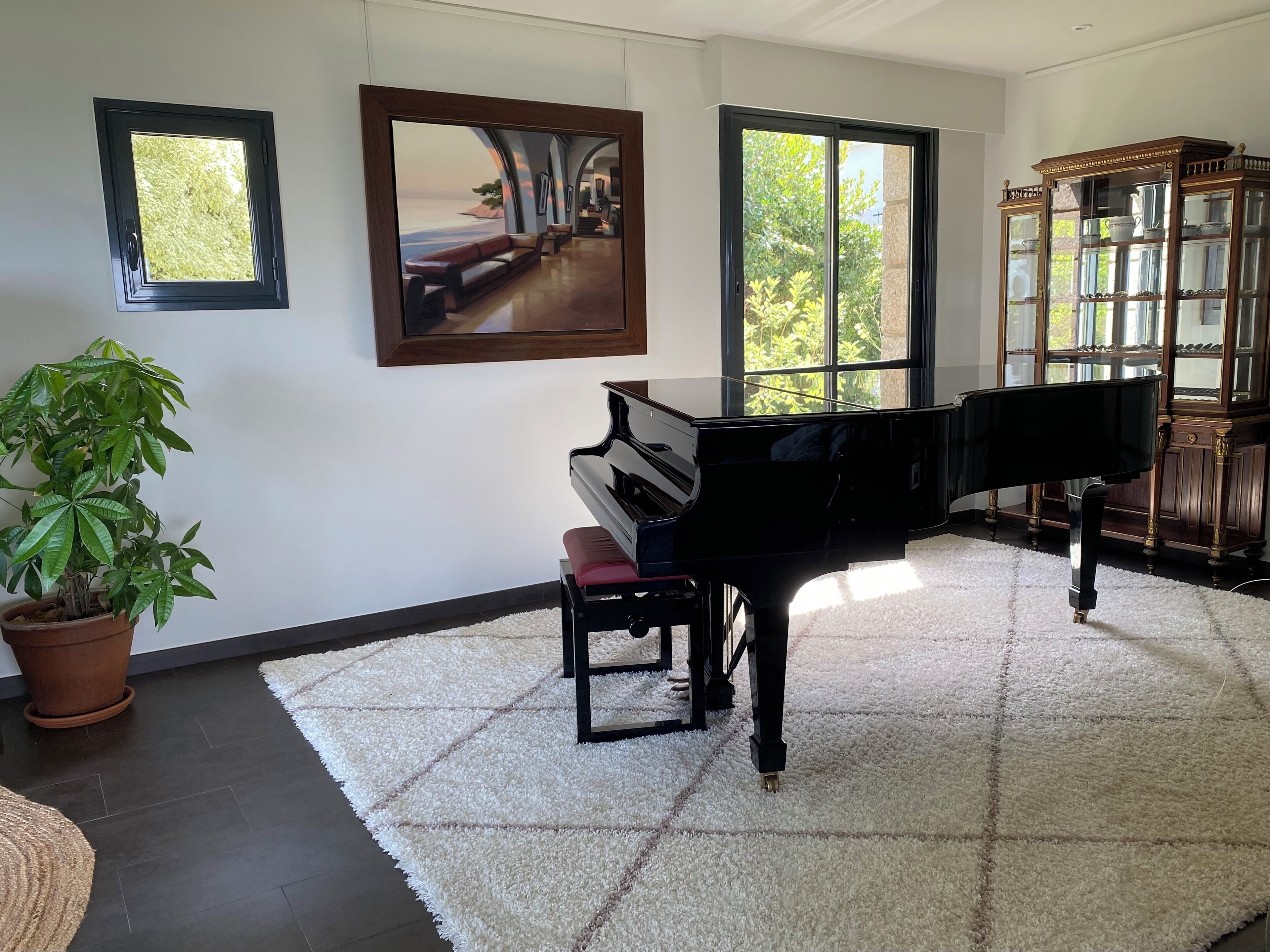 A room with a grand piano, a large window with a view, a potted plant, and a wooden cabinet.