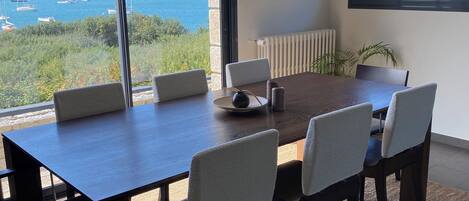 Un comedor moderno con una mesa grande y sillas, ventanas amplias que ofrecen vista al mar y barcos, y un radiador en la pared.