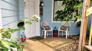 Eine Veranda mit einer weißen Tür, auf der „801“ steht, zwei weiße Stühle mit gemusterten Kissen, ein Vogelhaus und ein Blumentopf.