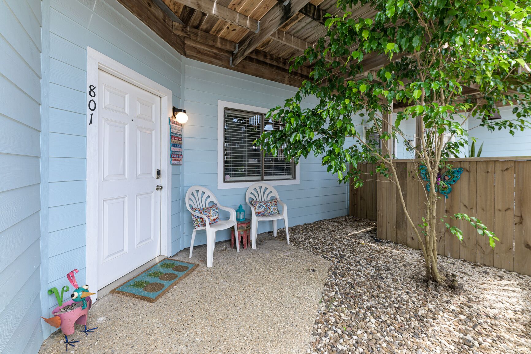 A house with a blue exterior, a white door marked "801", two white chairs with colorful cushions, a small tree, and a wooden fence.