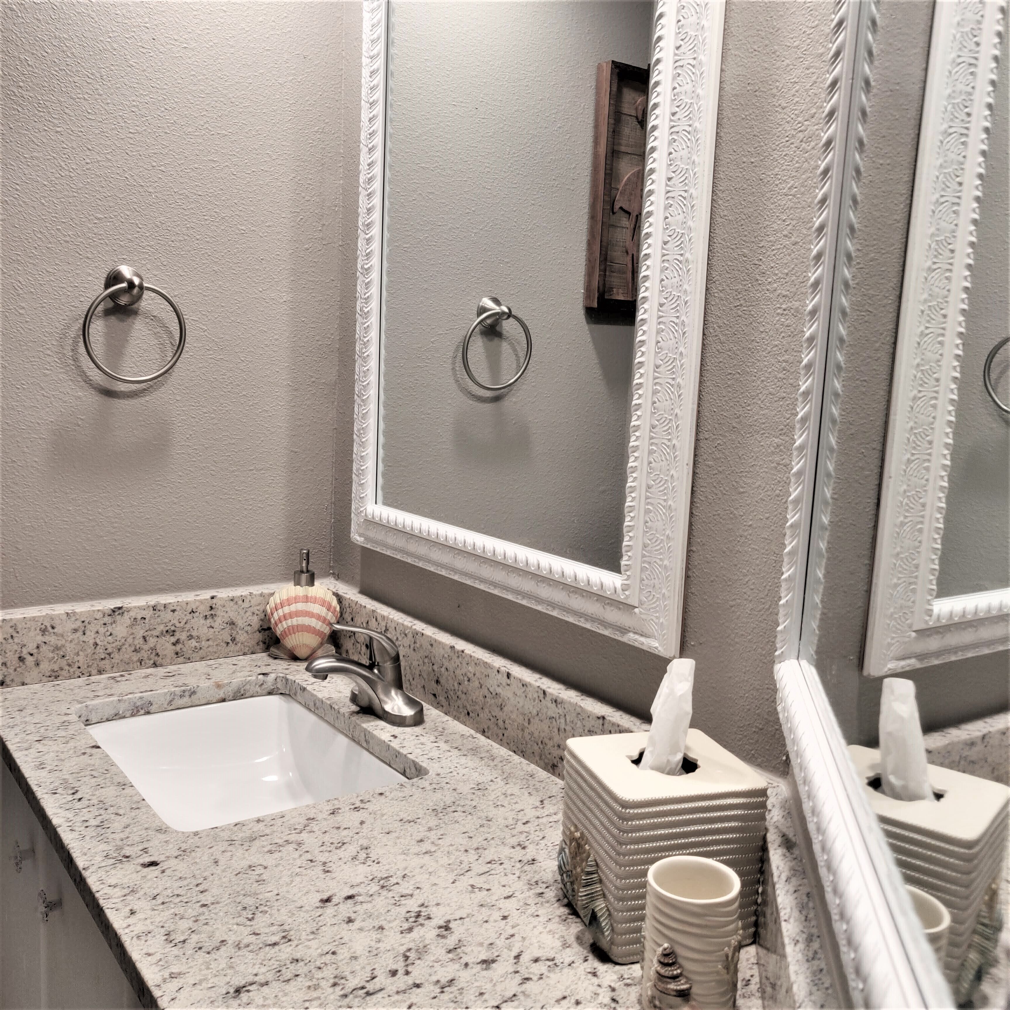 A bathroom with a granite countertop, a white sink, a mirror with ornate frame, and two towel rings.