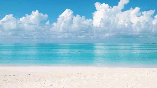 A beach with clear turquoise water and white sand under a blue sky with fluffy clouds.