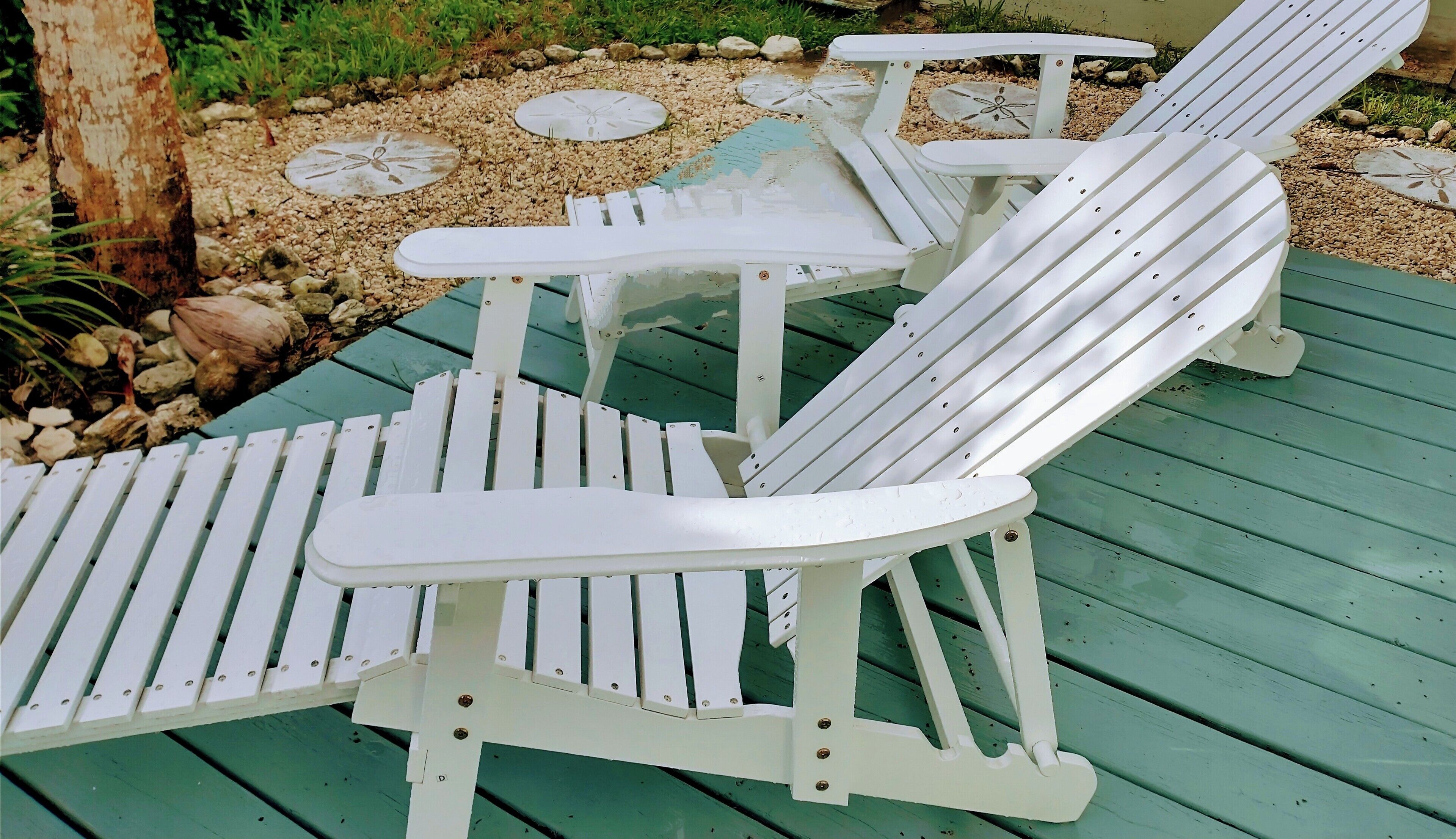 Un conjunto de sillas Adirondack blancas y una mesita pequeña en una terraza verde con borde de piedras.