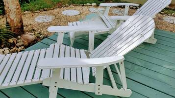 Un conjunto de sillas Adirondack blancas y una mesita pequeña en una terraza verde con borde de piedras.